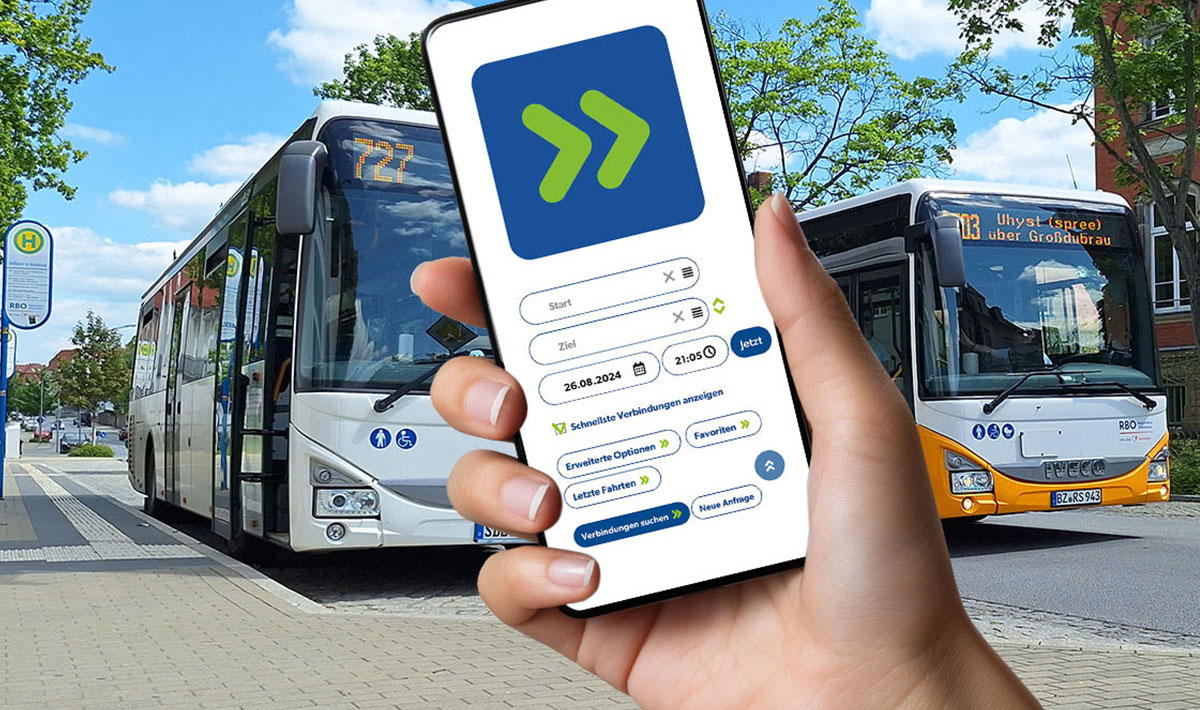 Hand hält ein Smartphone mit der PriMa+ÖV-App. Im Hintergrund stehen zwei Linienbusse an einer Bushaltestelle. Die App zeigt die Suche nach privaten Mitfahrmöglichkeiten und ÖPNV-Verbindungen.