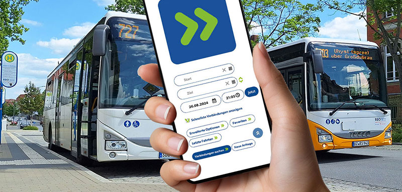 Hand hält ein Smartphone mit der PriMa+ÖV-App. Im Hintergrund stehen zwei Linienbusse an einer Bushaltestelle. Die App zeigt die Suche nach privaten Mitfahrmöglichkeiten und ÖPNV-Verbindungen.